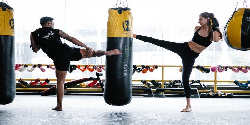 A man and woman kicking a heavy bag in the gym.