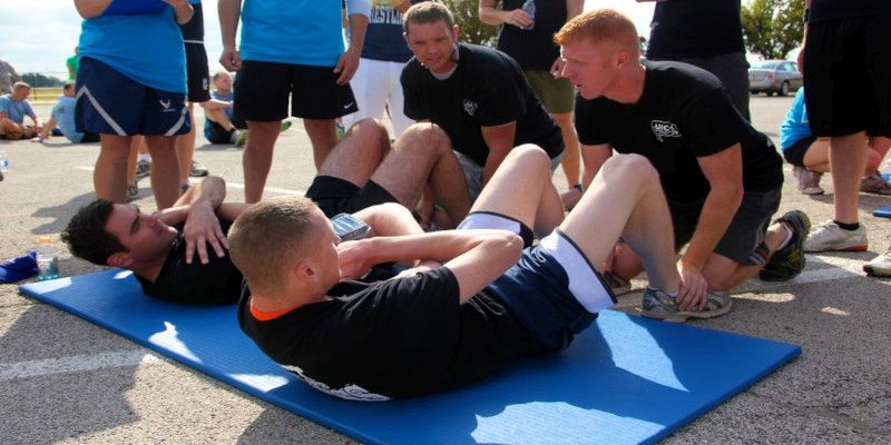 Picture of multiple men performing sit-ups outside on a blue mat with other people surrounding them.