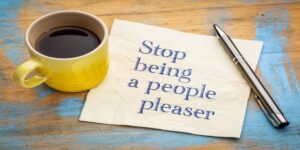 “Stop being a people pleaser” written down on a note with a cup of coffee and a pen next to it.