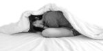 A woman lying in bed curled up suffering from anxiety and fear with the blanket covering her body.